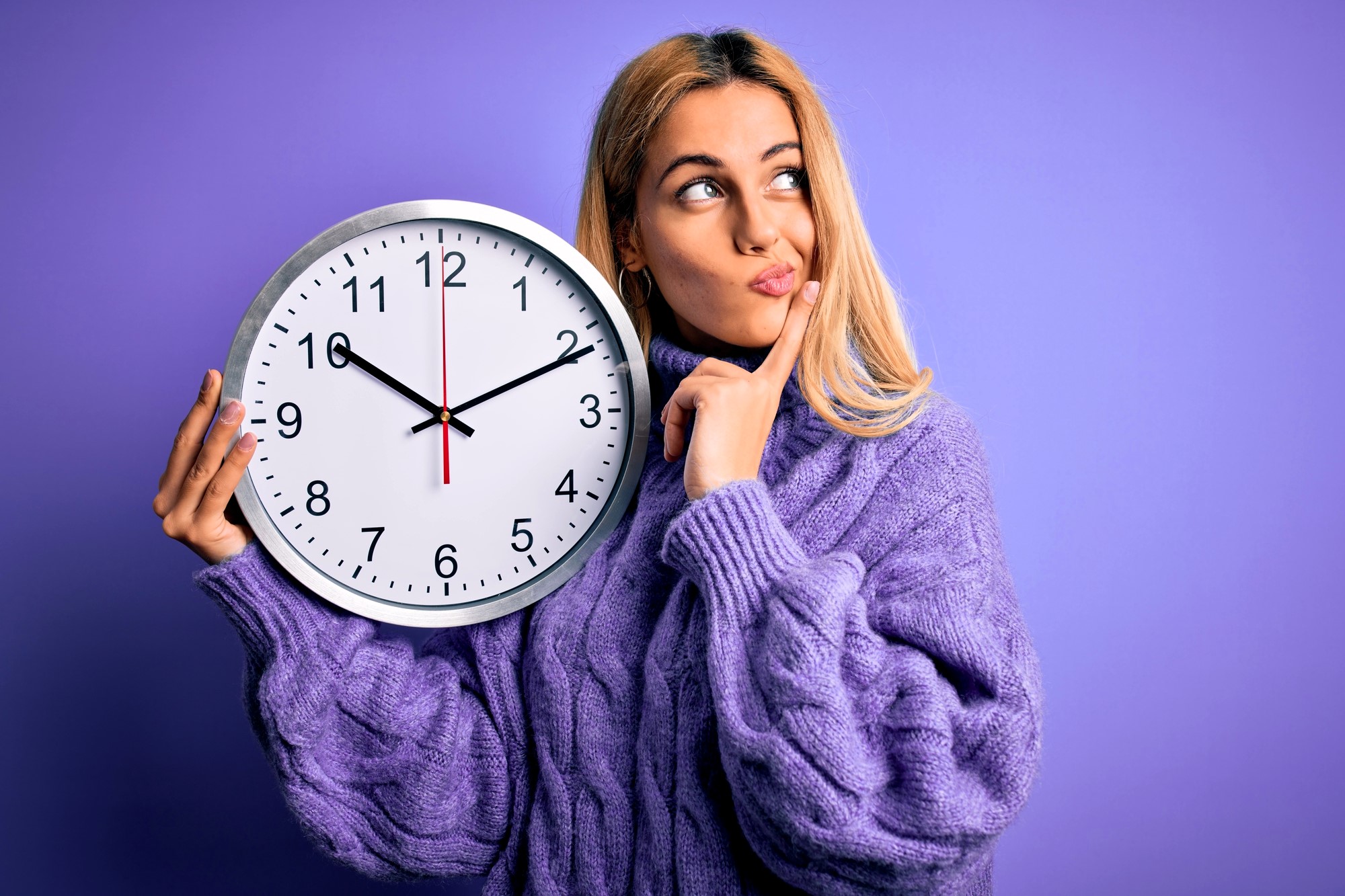 A person with long blonde hair, wearing a purple sweater, holds a large clock showing 8:20. They are looking upward with a thoughtful expression against a purple background.