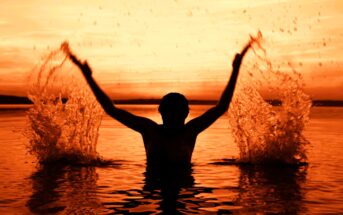 Silhouette of a person in water with arms raised, splashing droplets. The background is an intense orange sunset, reflecting on the rippling water surface.