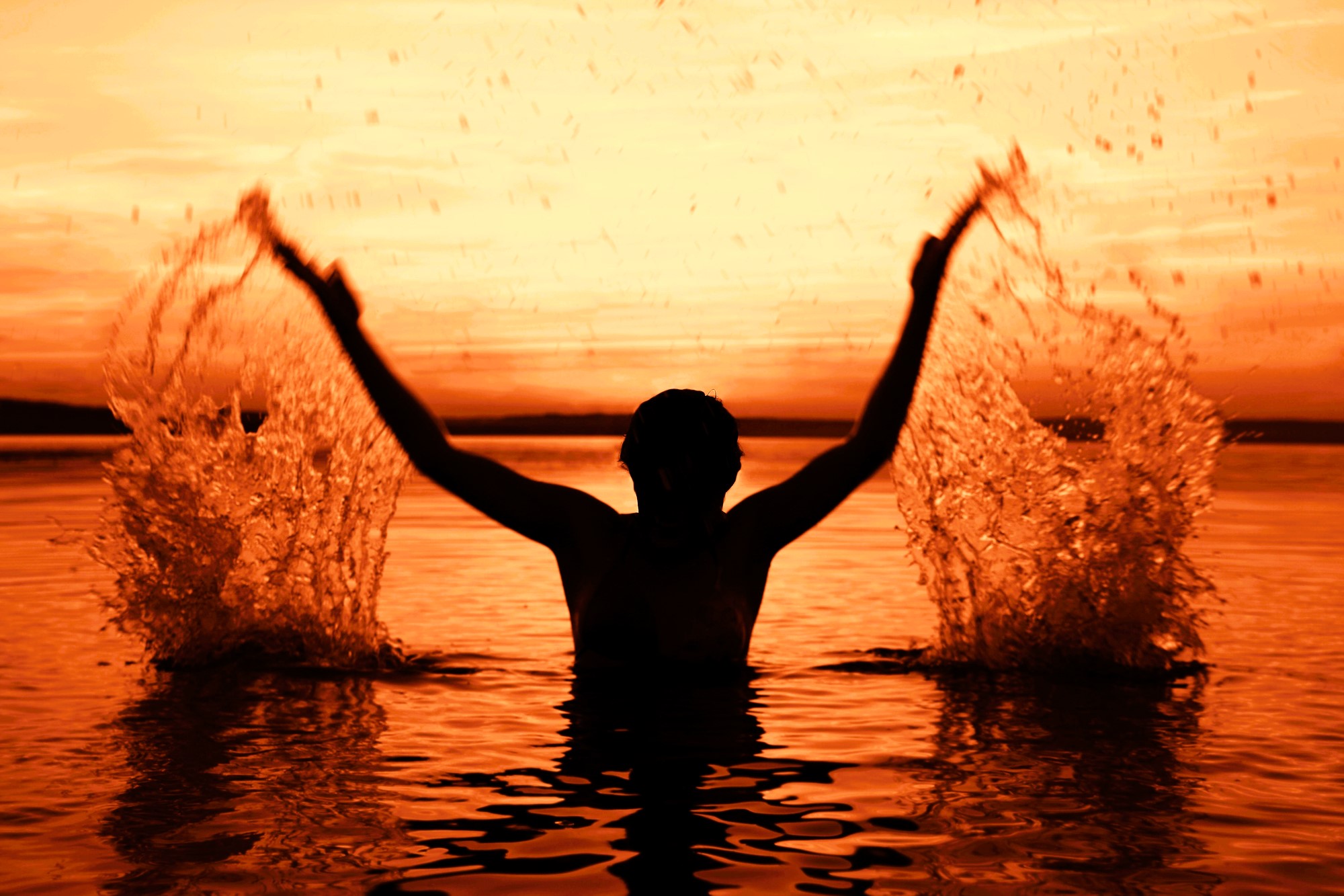 Silhouette of a person in water with arms raised, splashing droplets. The background is an intense orange sunset, reflecting on the rippling water surface.