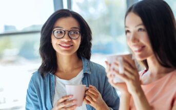 Two women enjoying coffee together, seated in a sunlit room. One is wearing glasses and a denim shirt, smiling, while the other holds a mug and looks content. Blurred background with large windows providing natural light.