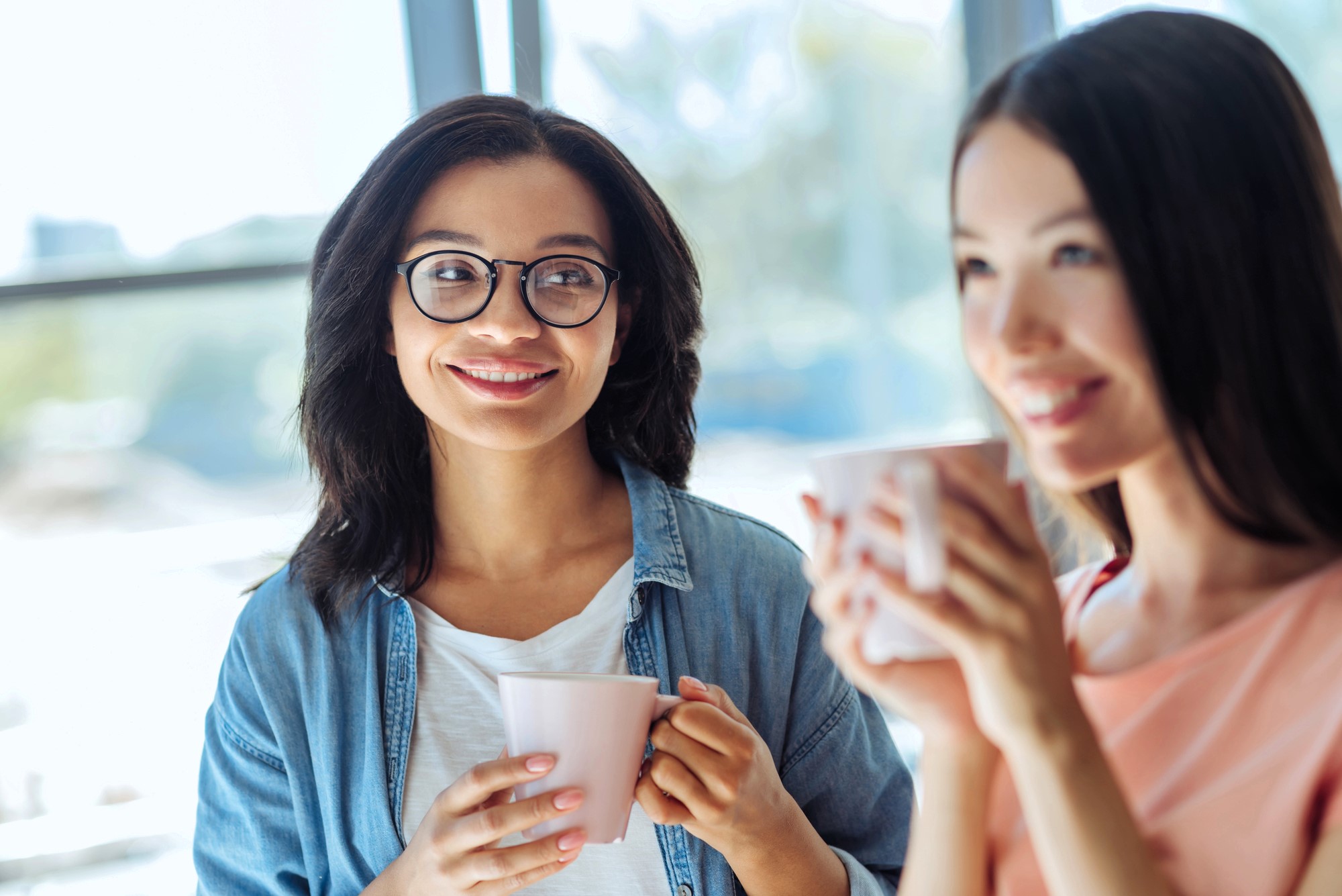 Two women enjoying coffee together, seated in a sunlit room. One is wearing glasses and a denim shirt, smiling, while the other holds a mug and looks content. Blurred background with large windows providing natural light.