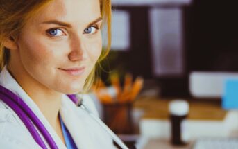 A person with auburn hair and blue eyes is wearing a white coat and a stethoscope, smiling slightly while sitting indoors. The background is blurred, showing a desk with office supplies and a computer.