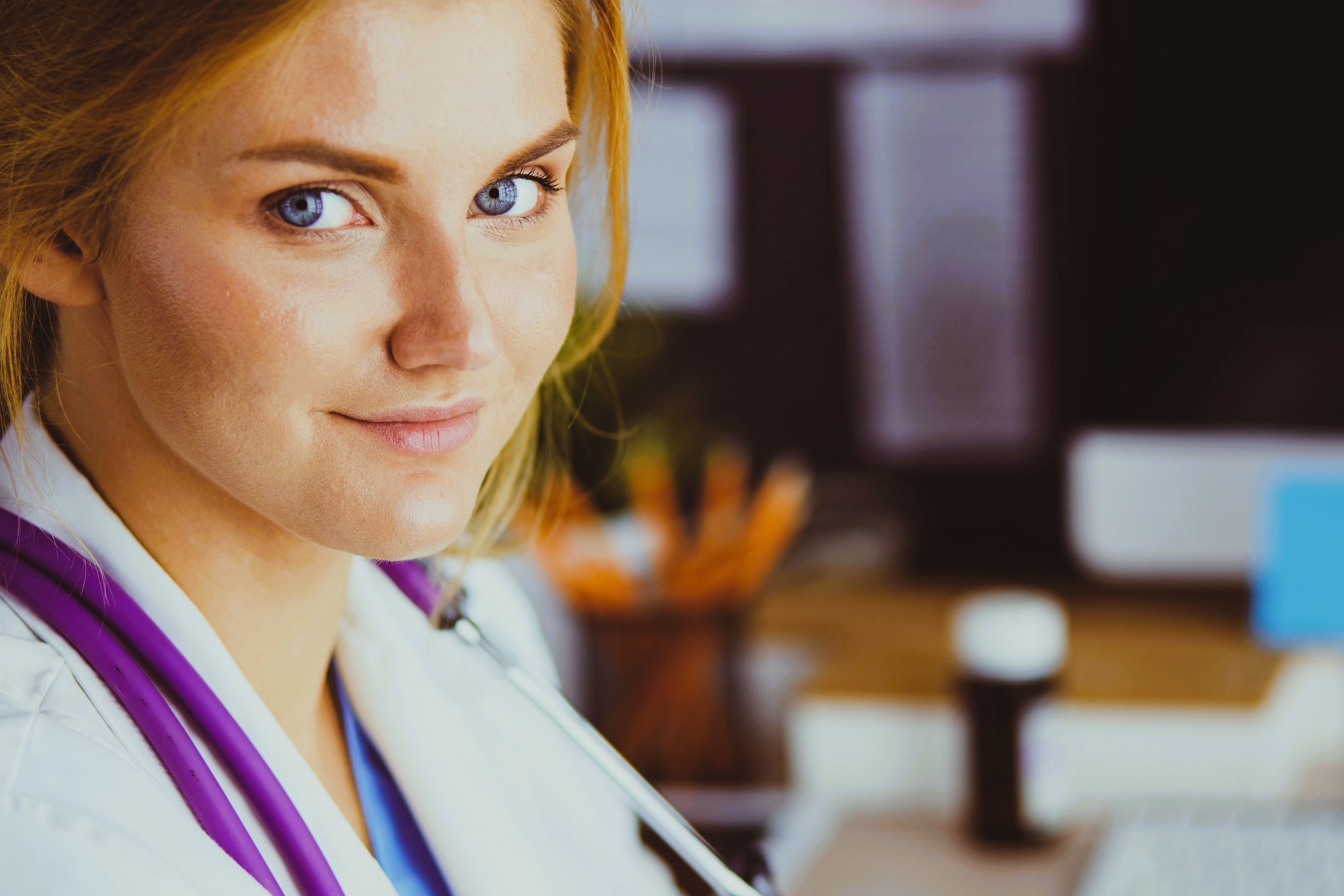 A person with auburn hair and blue eyes is wearing a white coat and a stethoscope, smiling slightly while sitting indoors. The background is blurred, showing a desk with office supplies and a computer.