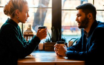 A woman and a man sit across from each other at a wooden table in a cafe, holding takeaway coffee cups. They are engaged in conversation, with a large window behind them showing a blurred outdoor scene.