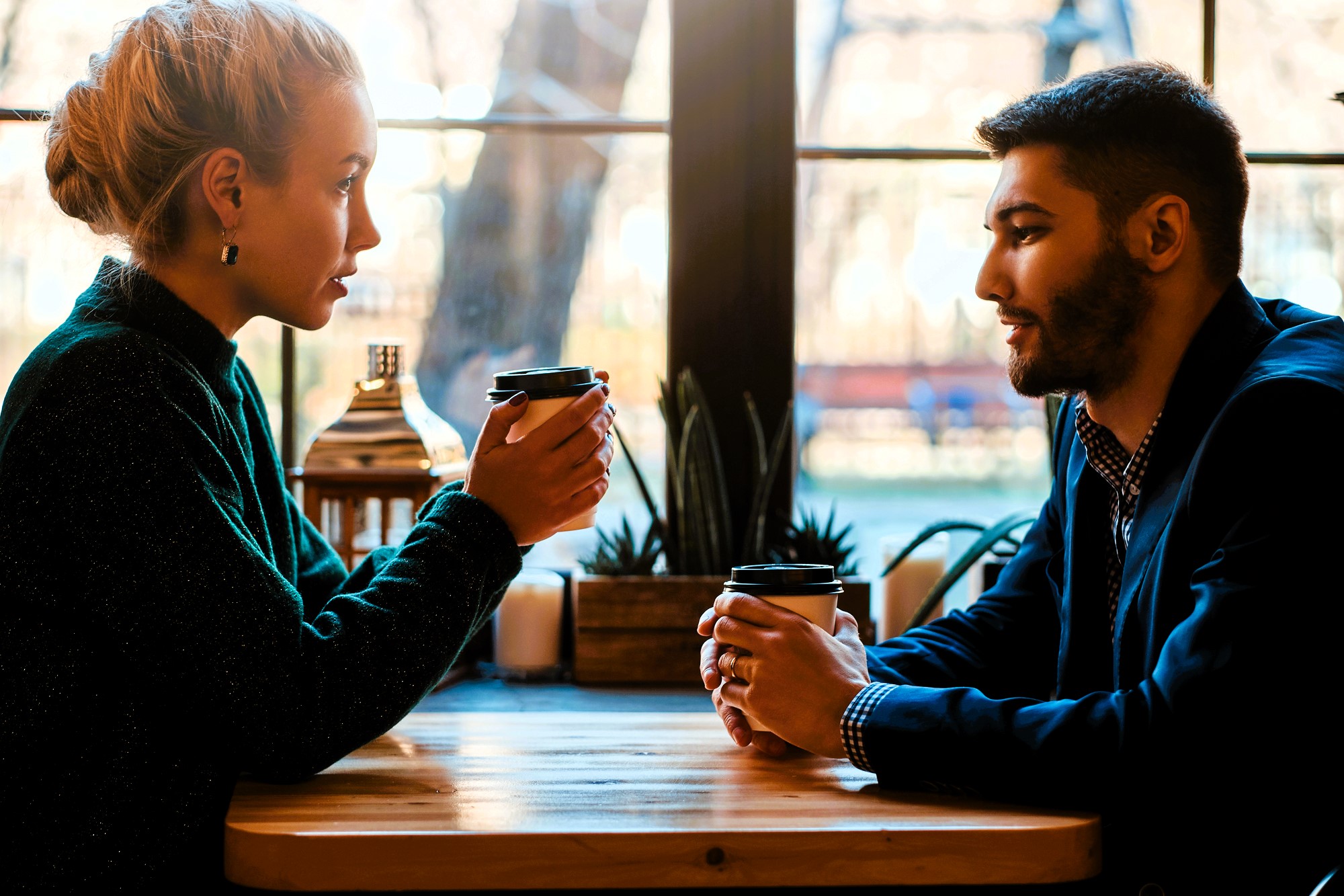 A woman and a man sit across from each other at a wooden table in a cafe, holding takeaway coffee cups. They are engaged in conversation, with a large window behind them showing a blurred outdoor scene.