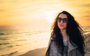 A woman with long dark hair wearing sunglasses smiles as she stands on a beach during sunset. The ocean waves are visible in the background, and the sky is a warm blend of yellow and orange hues.