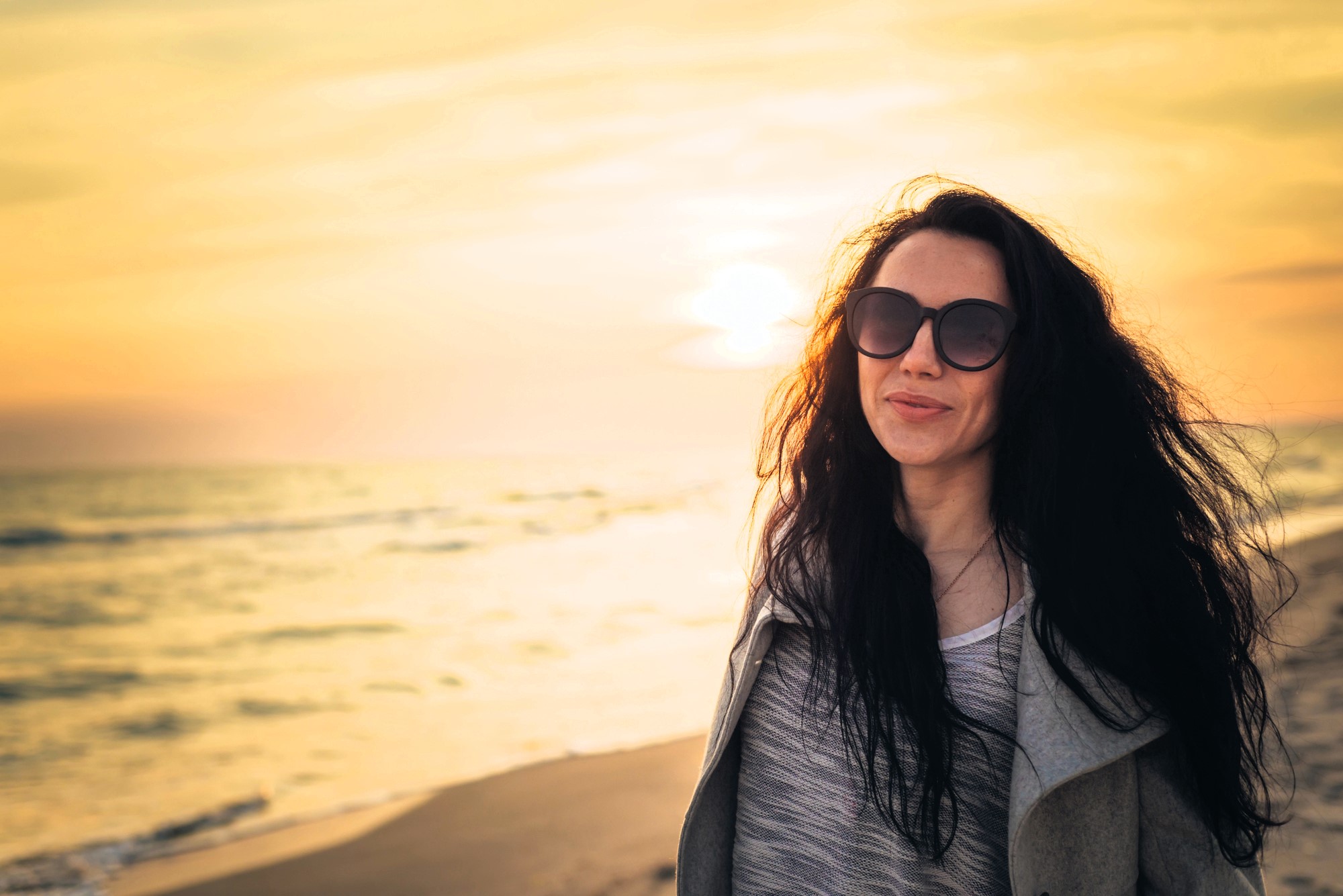 A woman with long dark hair wearing sunglasses smiles as she stands on a beach during sunset. The ocean waves are visible in the background, and the sky is a warm blend of yellow and orange hues.