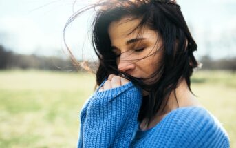 Woman in a blue sweater stands outdoors with eyes closed and hair blowing in the wind. She gently bites or holds the sweater near her mouth. The background is a soft-focus landscape, suggesting a serene, peaceful setting.