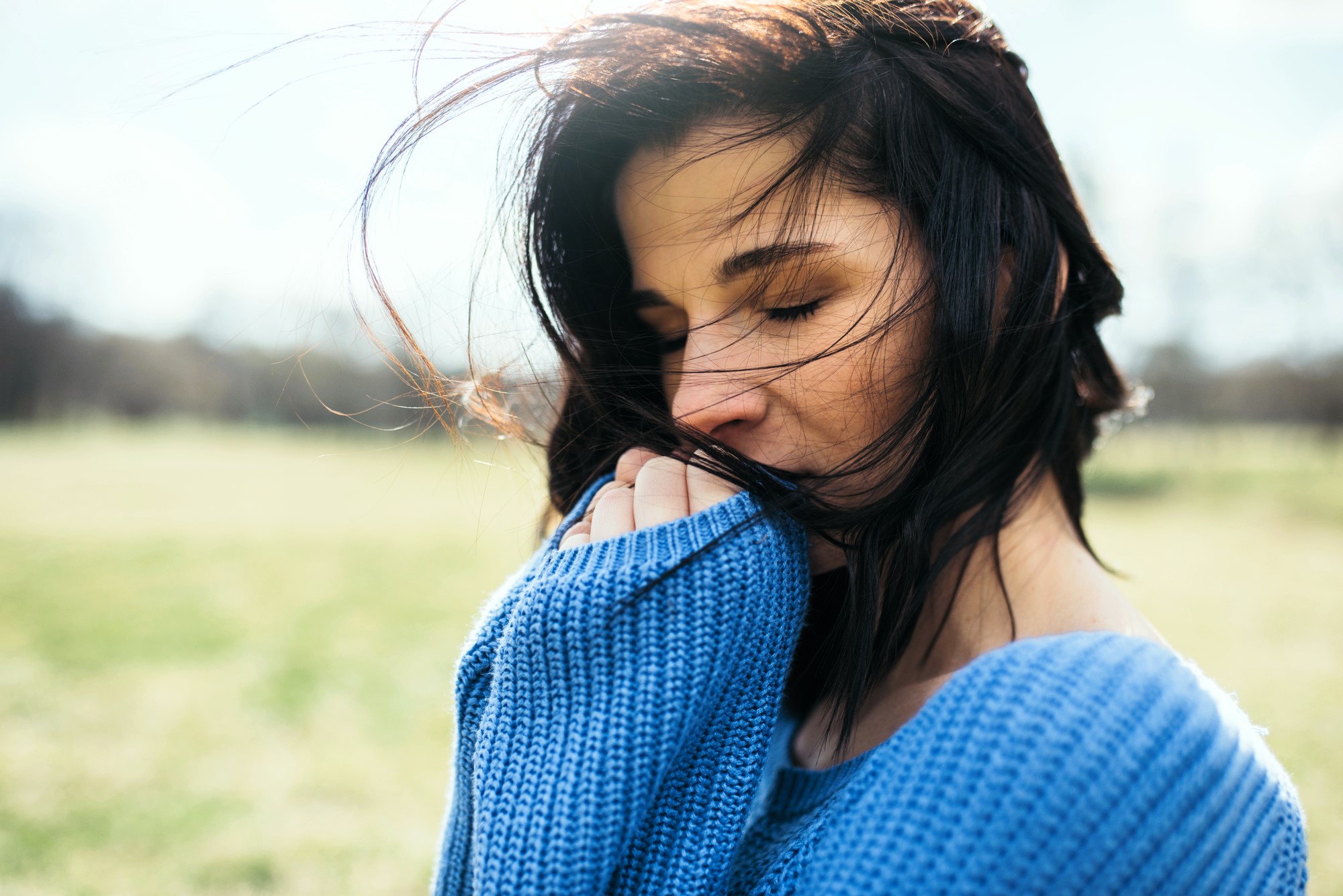 Woman in a blue sweater stands outdoors with eyes closed and hair blowing in the wind. She gently bites or holds the sweater near her mouth. The background is a soft-focus landscape, suggesting a serene, peaceful setting.