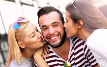 A bearded man in a striped shirt smiles as two women kiss him on the cheeks. One woman has blonde hair and wears sunglasses, while the other has long brown hair. They are outdoors, with a soft-focus city background.