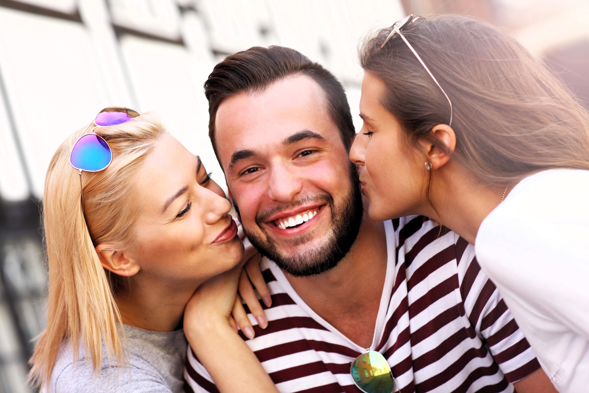 A bearded man in a striped shirt smiles as two women kiss him on the cheeks. One woman has blonde hair and wears sunglasses, while the other has long brown hair. They are outdoors, with a soft-focus city background.