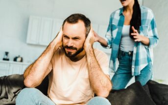 A man sits covering his ears, appearing frustrated. A woman stands nearby, gesturing as if speaking. They are in a room with a modern, light-colored interior. The man wears a beige t-shirt, and the woman is in a blue checkered shirt and jeans.