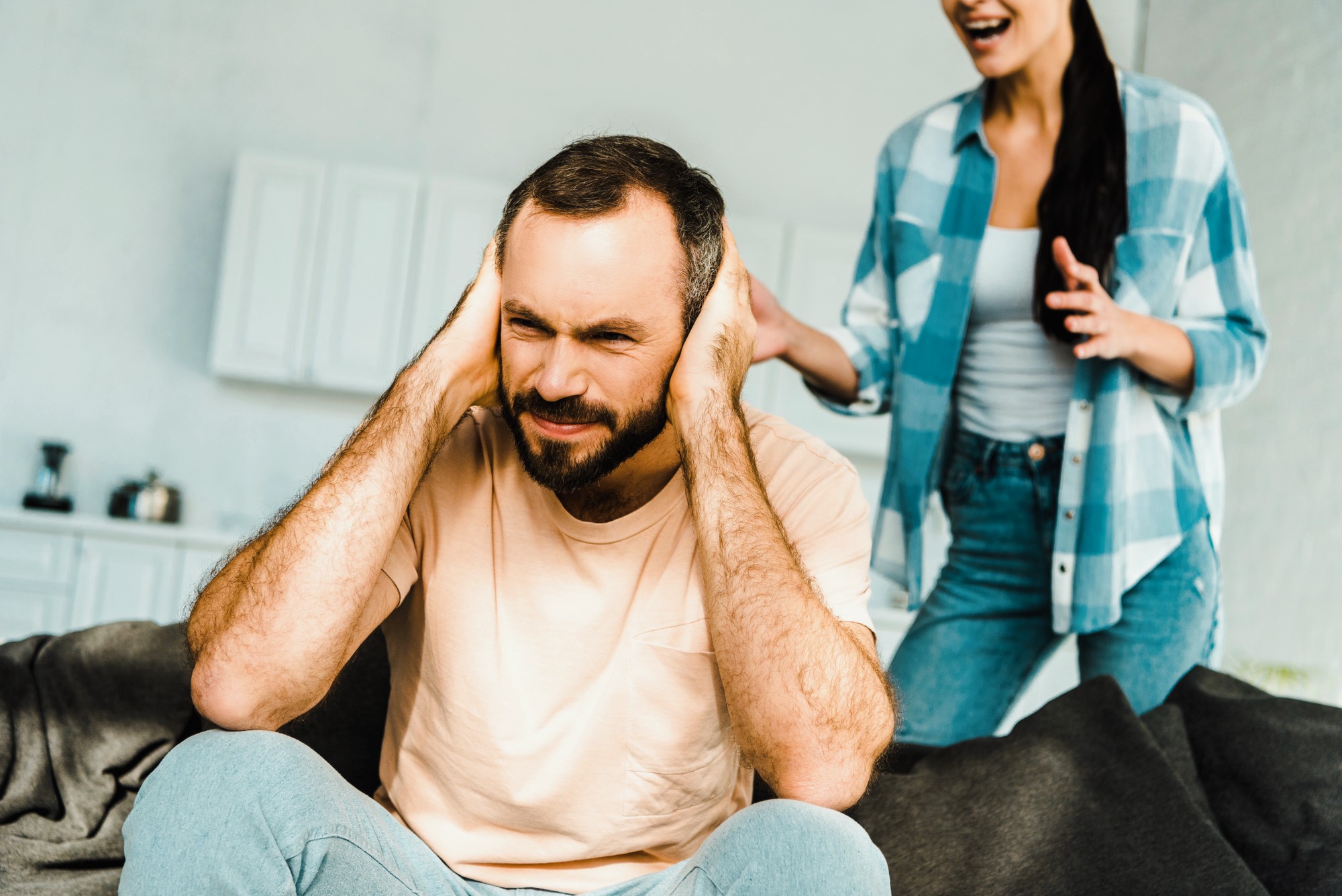 A man sits covering his ears, appearing frustrated. A woman stands nearby, gesturing as if speaking. They are in a room with a modern, light-colored interior. The man wears a beige t-shirt, and the woman is in a blue checkered shirt and jeans.