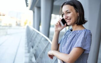 A woman with short hair and a checkered top is smiling while talking on a smartphone. She is leaning against a railing, standing in an outdoor setting with modern architecture in the background.