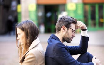 A man and woman sit back to back outdoors, both looking upset. The man, wearing glasses and a suit, rests his head on his hand. The woman, in a tan coat, has her arms crossed. They are in a public space with green and orange storefronts in the background.