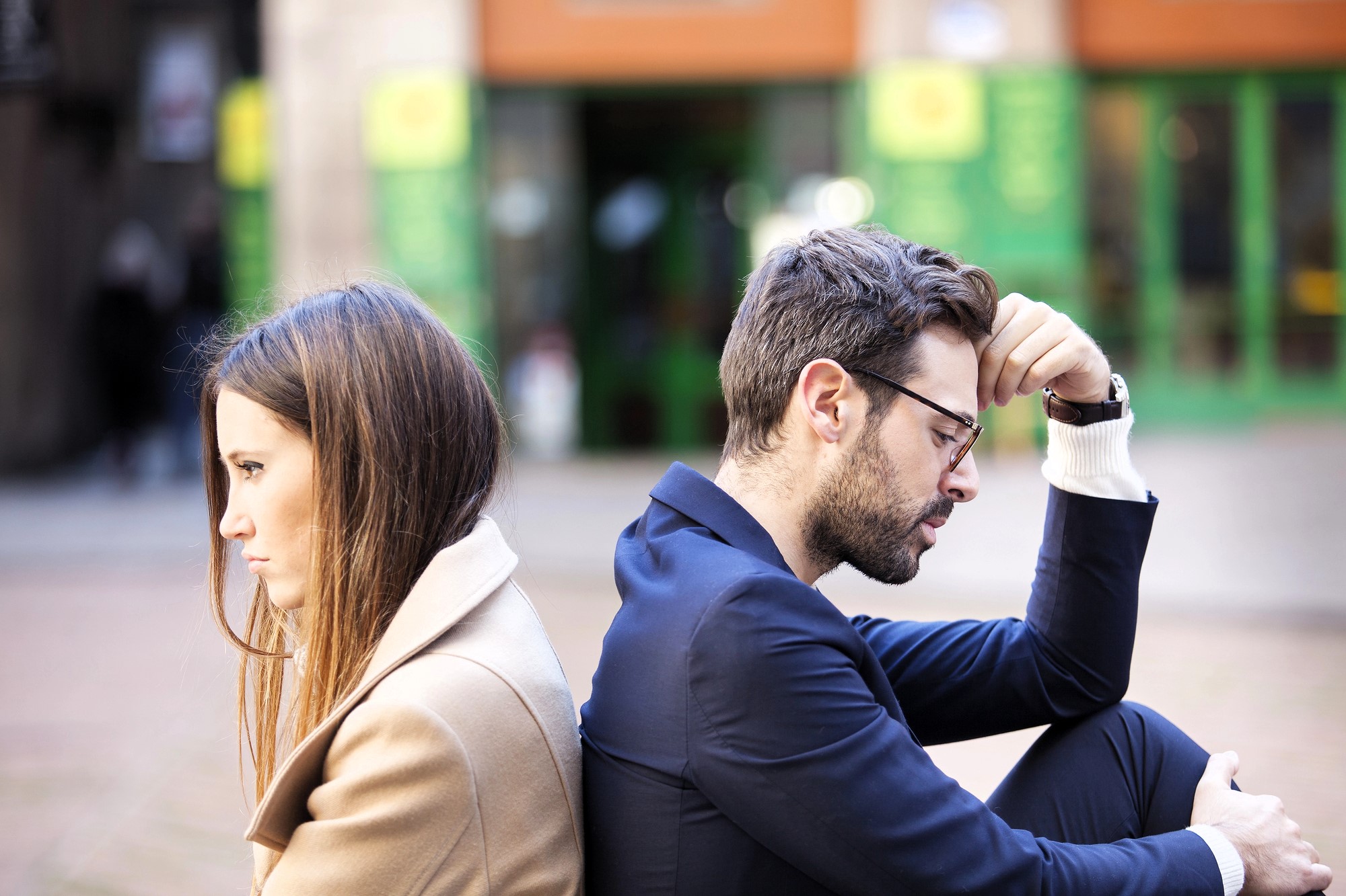 A man and woman sit back to back outdoors, both looking upset. The man, wearing glasses and a suit, rests his head on his hand. The woman, in a tan coat, has her arms crossed. They are in a public space with green and orange storefronts in the background.