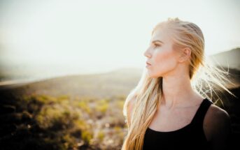 A woman with long blonde hair stands in a sunlit field, looking to the right. She is wearing a black tank top. The background is a blurred, sunny landscape of grass and hills.