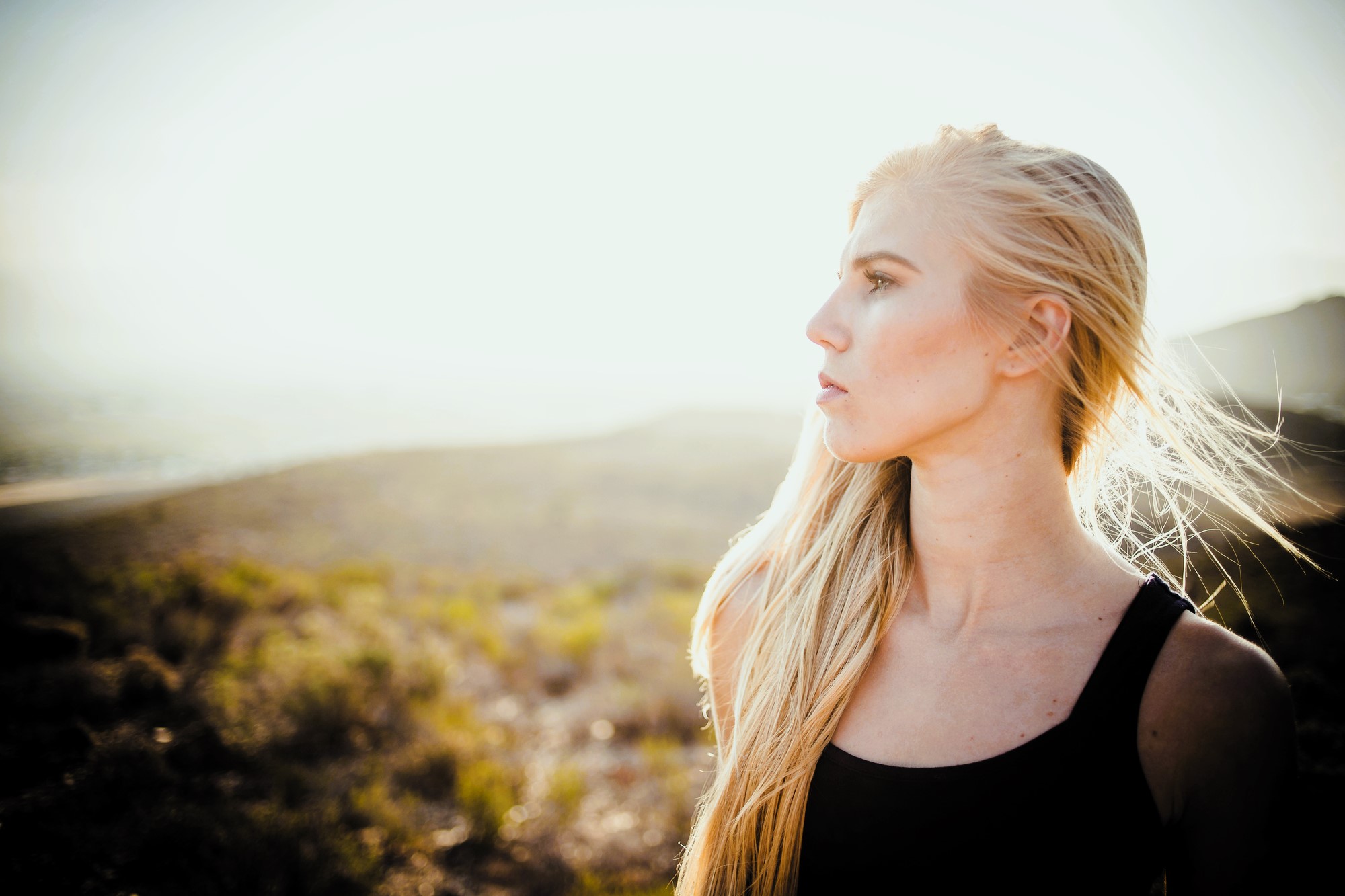 A woman with long blonde hair stands in a sunlit field, looking to the right. She is wearing a black tank top. The background is a blurred, sunny landscape of grass and hills.