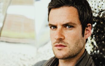 Man with short dark hair and stubble looks intensely at the camera. He's wearing a gray scarf, and the background is a blurred mix of light colors, possibly metallic or reflective surfaces.