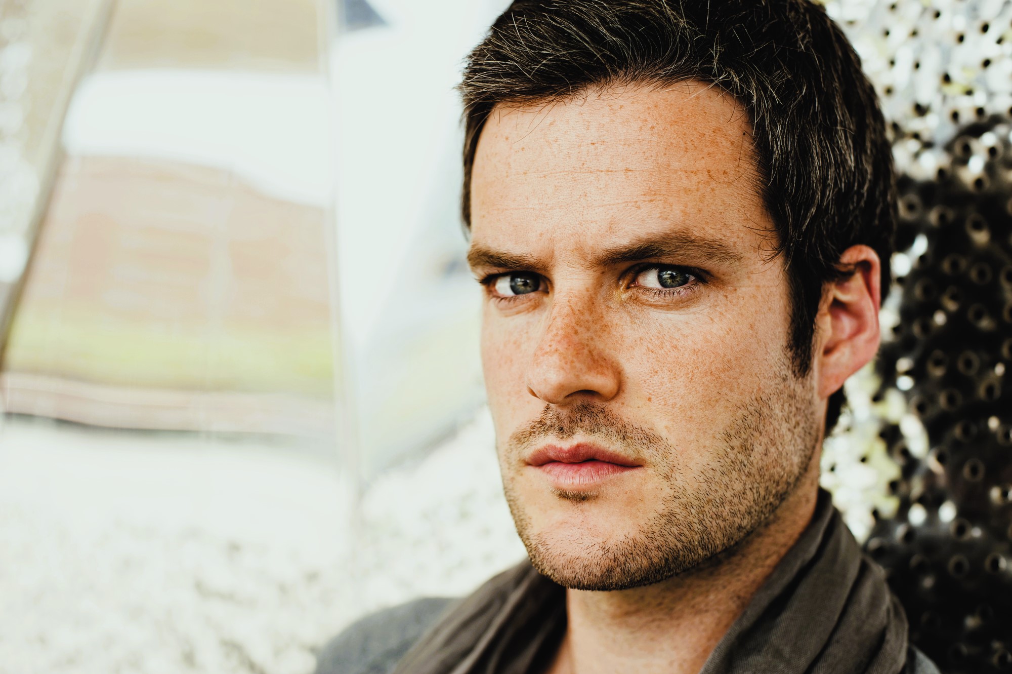 Man with short dark hair and stubble looks intensely at the camera. He's wearing a gray scarf, and the background is a blurred mix of light colors, possibly metallic or reflective surfaces.