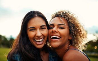 Two women smiling widely, leaning their heads together outdoors. One has long dark hair, the other has curly blonde hair. They're both happy and relaxed, with greenery and a bright sky in the background.