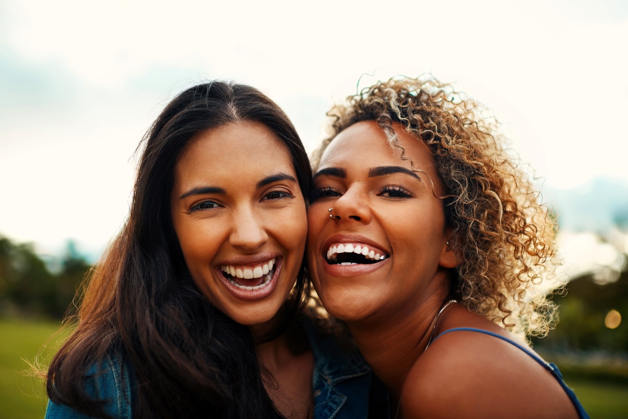 Two women smiling widely, leaning their heads together outdoors. One has long dark hair, the other has curly blonde hair. They're both happy and relaxed, with greenery and a bright sky in the background.