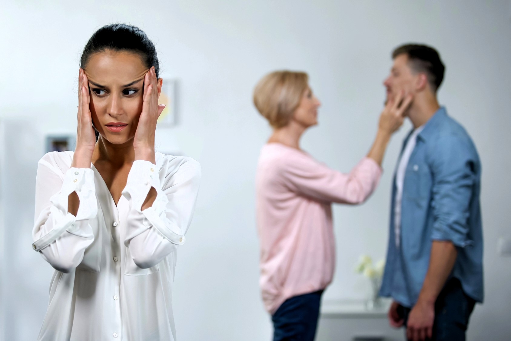 A woman in the foreground looks worried, holding her head with both hands. In the background, a couple is experiencing an argument; a woman is gesturing towards a man's face. The scene suggests tension among the three people.