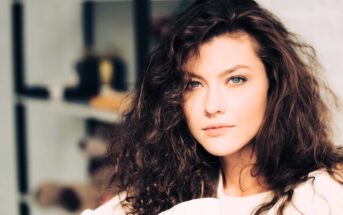 A woman with long, curly brown hair and blue eyes gazes intently at the camera. She is indoors, with a soft-focus bookshelf in the background. The lighting casts a warm glow on her face.