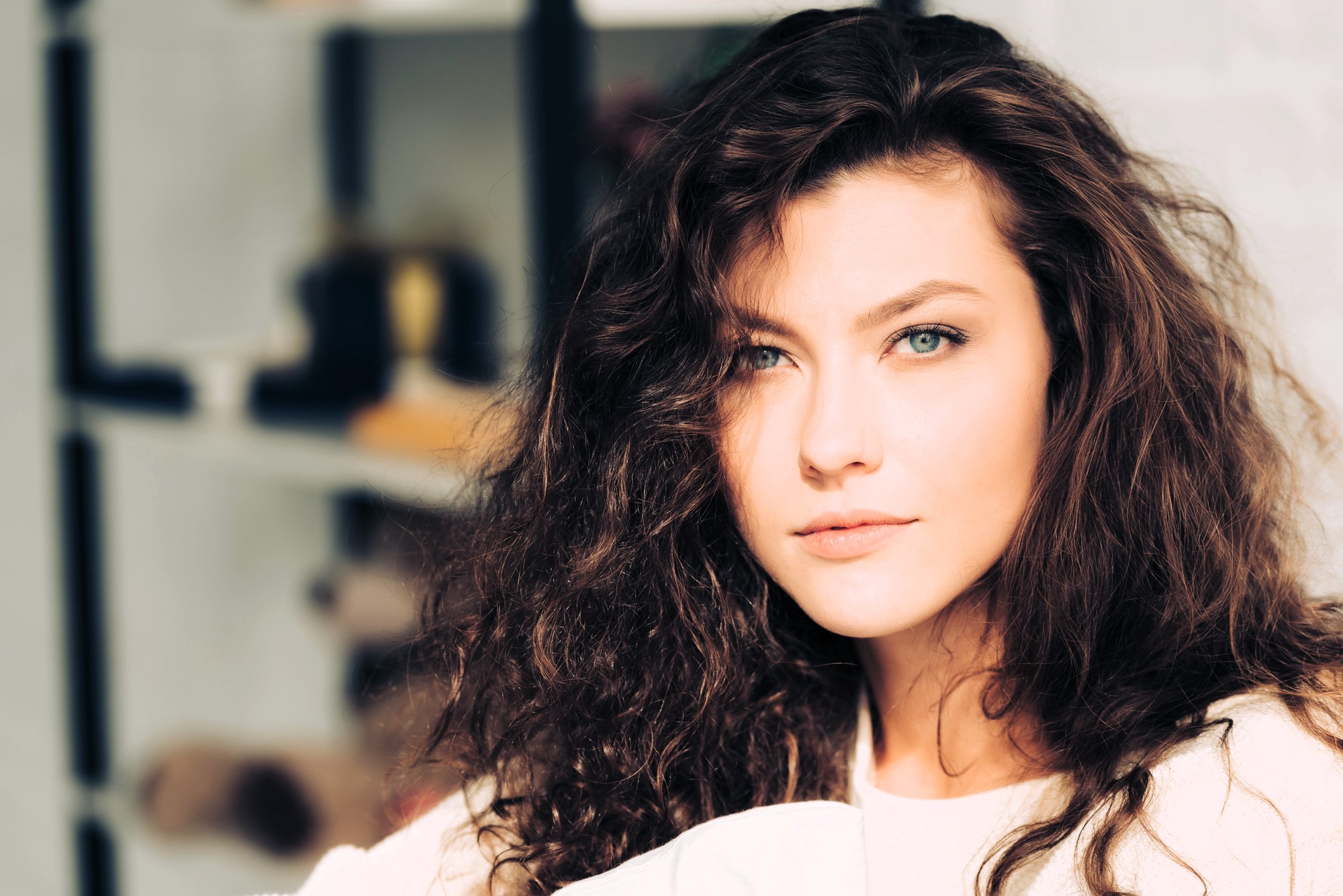 A woman with long, curly brown hair and blue eyes gazes intently at the camera. She is indoors, with a soft-focus bookshelf in the background. The lighting casts a warm glow on her face.