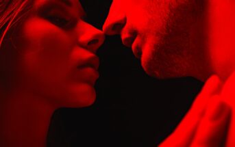 Close-up of two people in an intimate pose, their faces lit by red lighting against a dark background. They are facing each other, capturing a moment of anticipation or connection.