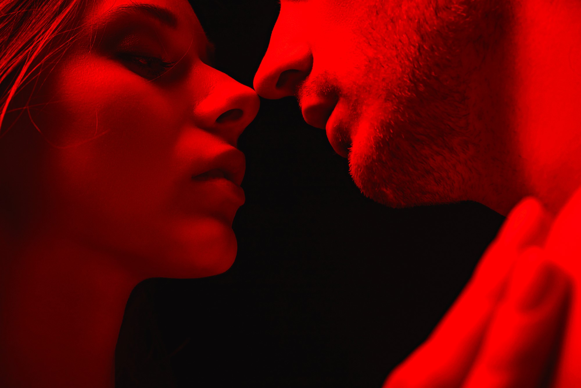 Close-up of two people in an intimate pose, their faces lit by red lighting against a dark background. They are facing each other, capturing a moment of anticipation or connection.