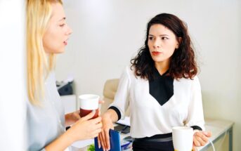 Two women standing in an office exchanging cups of coffee. One is leaning against a desk while the other speaks animatedly. They appear to be having a thoughtful conversation. Papers and office supplies are visible in the background.