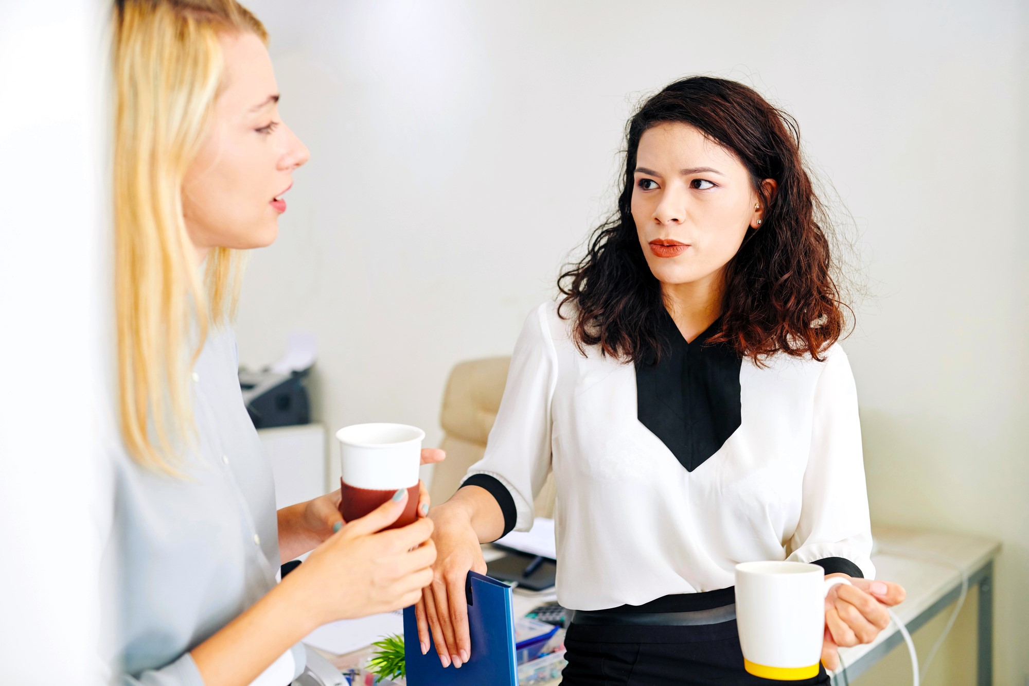 Two women standing in an office exchanging cups of coffee. One is leaning against a desk while the other speaks animatedly. They appear to be having a thoughtful conversation. Papers and office supplies are visible in the background.