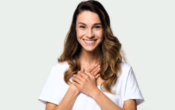 A person with long brown hair smiles warmly, hands clasped together at the chest. They wear a white T-shirt against a light grey background.