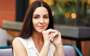 A woman with long dark hair and light skin is sitting outdoors in front of a blurred background with warm lights. She is resting her chin on her hands and wearing a sleeveless white top, looking at the camera with a gentle smile.