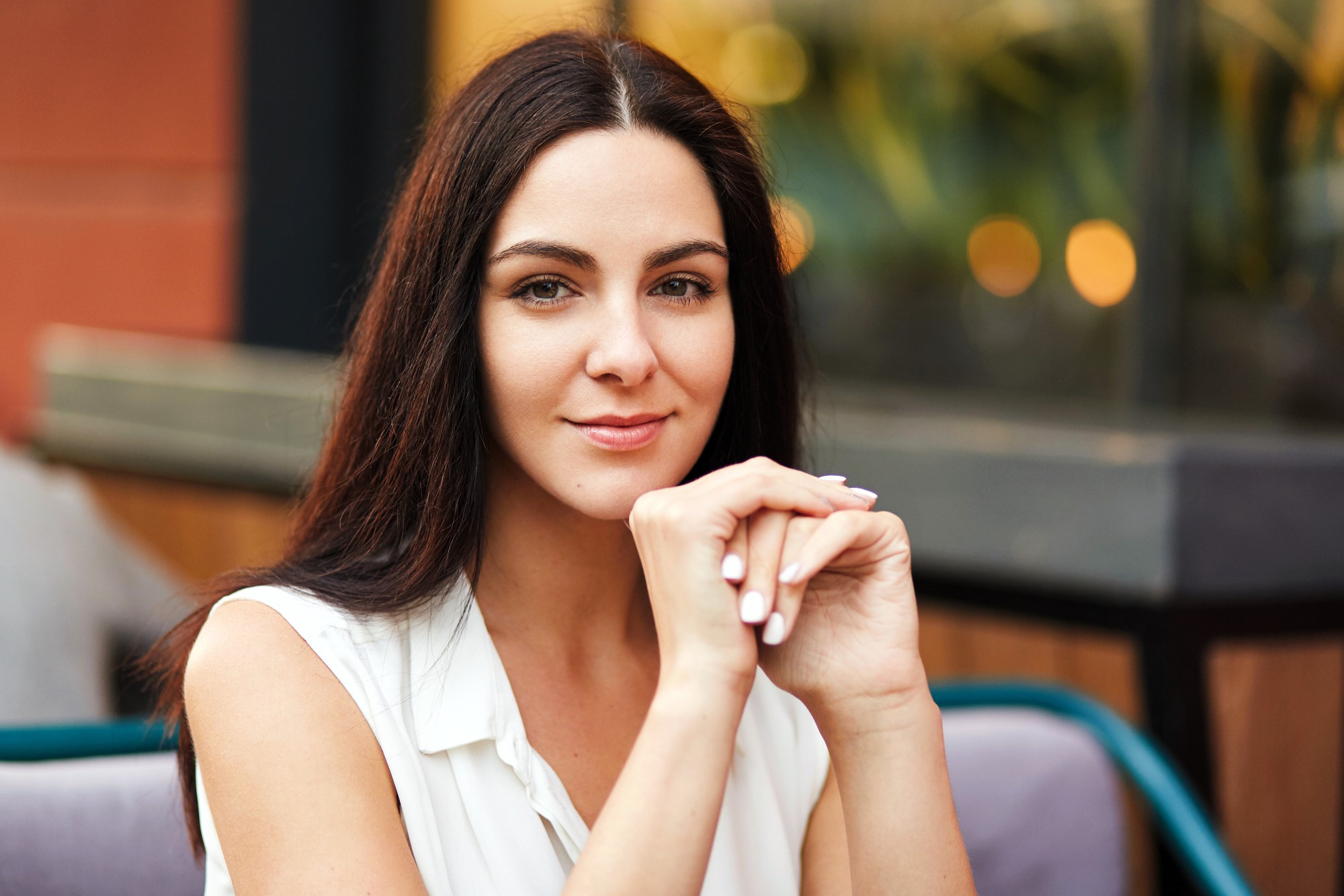 A woman with long dark hair and light skin is sitting outdoors in front of a blurred background with warm lights. She is resting her chin on her hands and wearing a sleeveless white top, looking at the camera with a gentle smile.