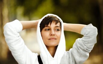 A person with short hair wearing a white hoodie stands outdoors with hands behind their head, smiling slightly. The background is blurred green foliage, conveying a relaxed and serene atmosphere.