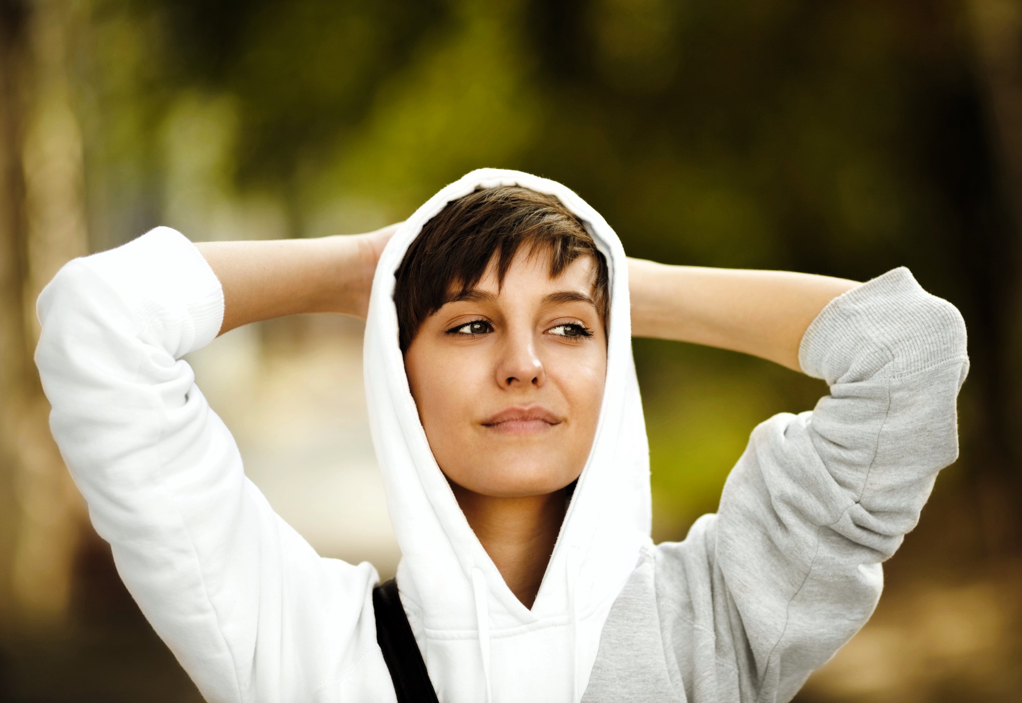 A person with short hair wearing a white hoodie stands outdoors with hands behind their head, smiling slightly. The background is blurred green foliage, conveying a relaxed and serene atmosphere.