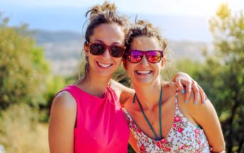 Two women smiling and wearing sunglasses stand together outdoors. One wears a bright pink top, and the other a floral dress. Sunlight filters through the trees, creating a warm atmosphere with a scenic view in the background.