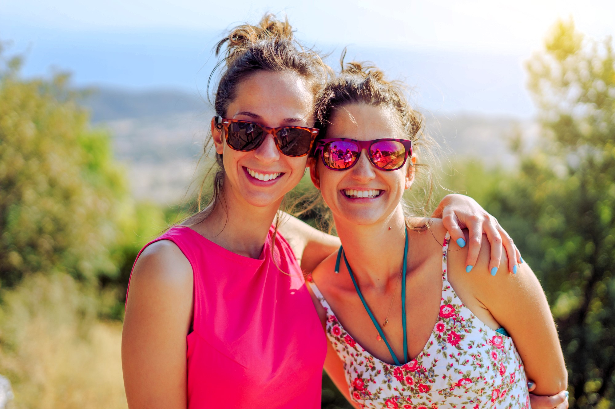 Two women smiling and wearing sunglasses stand together outdoors. One wears a bright pink top, and the other a floral dress. Sunlight filters through the trees, creating a warm atmosphere with a scenic view in the background.