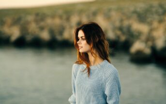 A woman with long brown hair stands by a body of water, wearing a light blue sweater. The background is blurred with rocks and grass, suggesting a serene outdoor setting. She gazes into the distance with a calm expression.