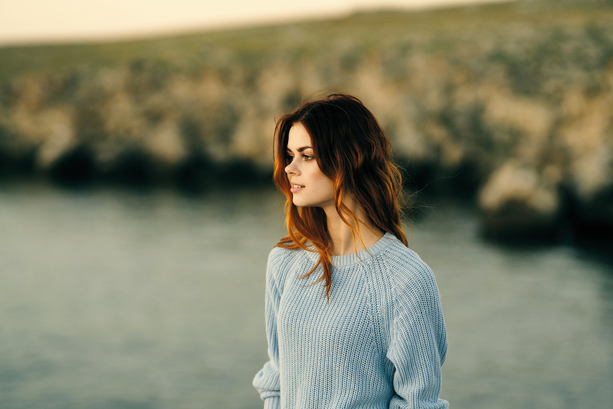 A woman with long brown hair stands by a body of water, wearing a light blue sweater. The background is blurred with rocks and grass, suggesting a serene outdoor setting. She gazes into the distance with a calm expression.