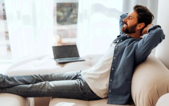 A man in a blue shirt and jeans is relaxing on a beige sofa with his hands behind his head, eyes closed. A laptop sits open beside him. Sunlight filters through sheer curtains in the background.