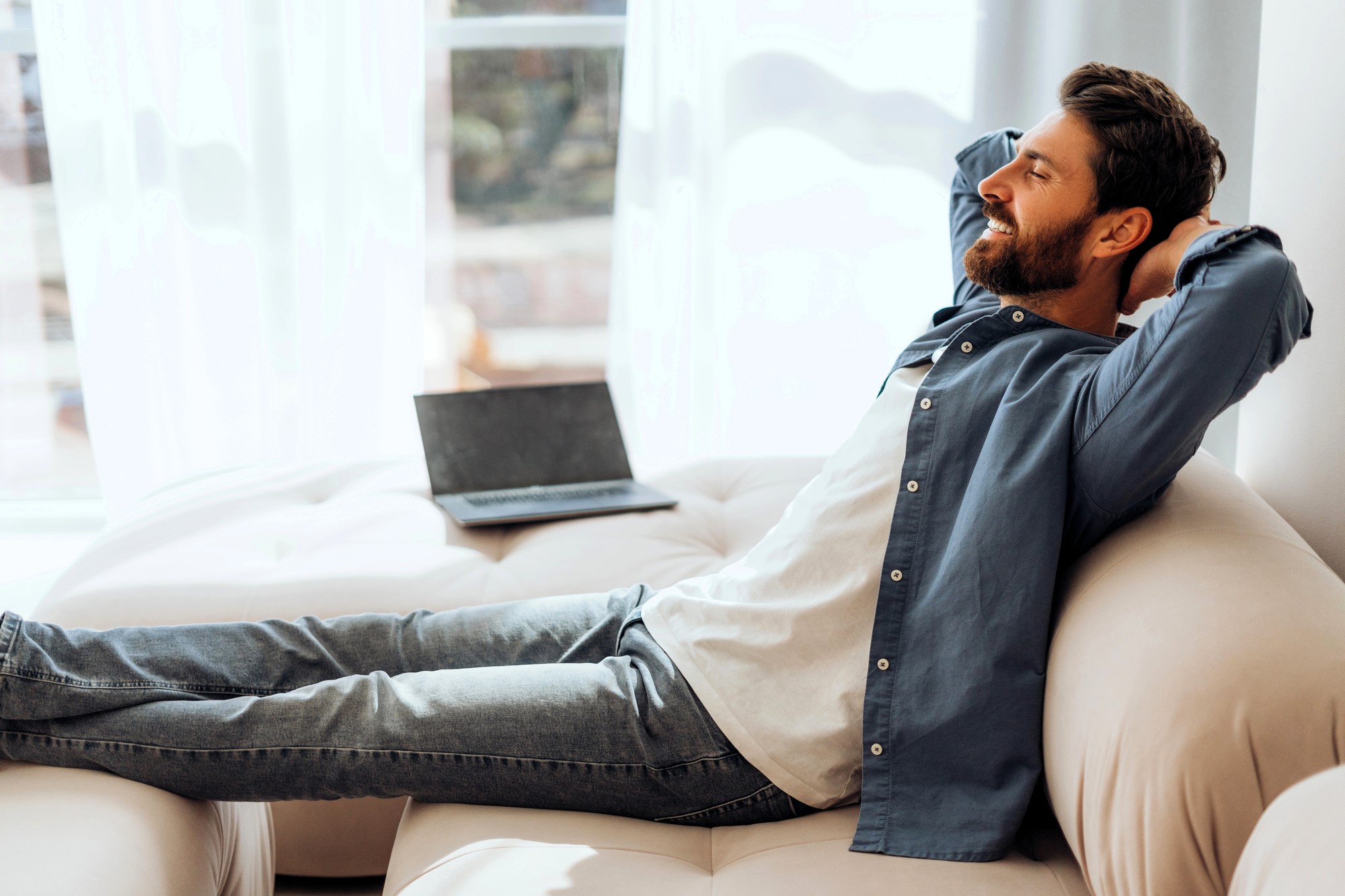 A man in a blue shirt and jeans is relaxing on a beige sofa with his hands behind his head, eyes closed. A laptop sits open beside him. Sunlight filters through sheer curtains in the background.