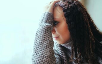 A woman with curly hair, wearing a gray sweater, rests her head on her hand, gazing pensively out of a window. The background is softly blurred, giving a serene and contemplative ambiance.