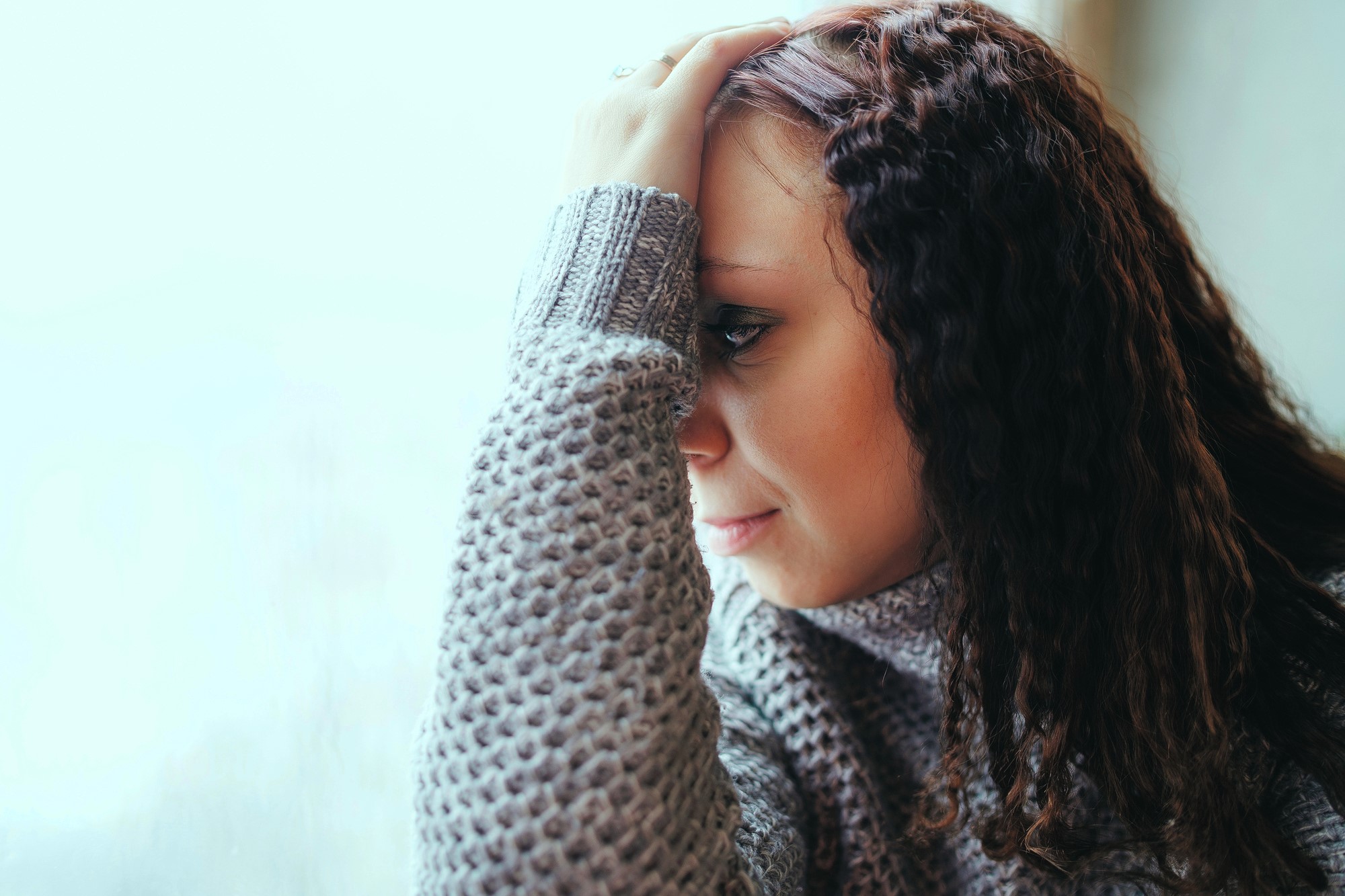 A woman with curly hair, wearing a gray sweater, rests her head on her hand, gazing pensively out of a window. The background is softly blurred, giving a serene and contemplative ambiance.