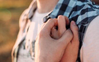 Close-up of two intertwined hands. One hand holds the other's shoulder, over a blue and white checkered shirt, signifying support or connection. The background is softly blurred with warm, natural lighting.