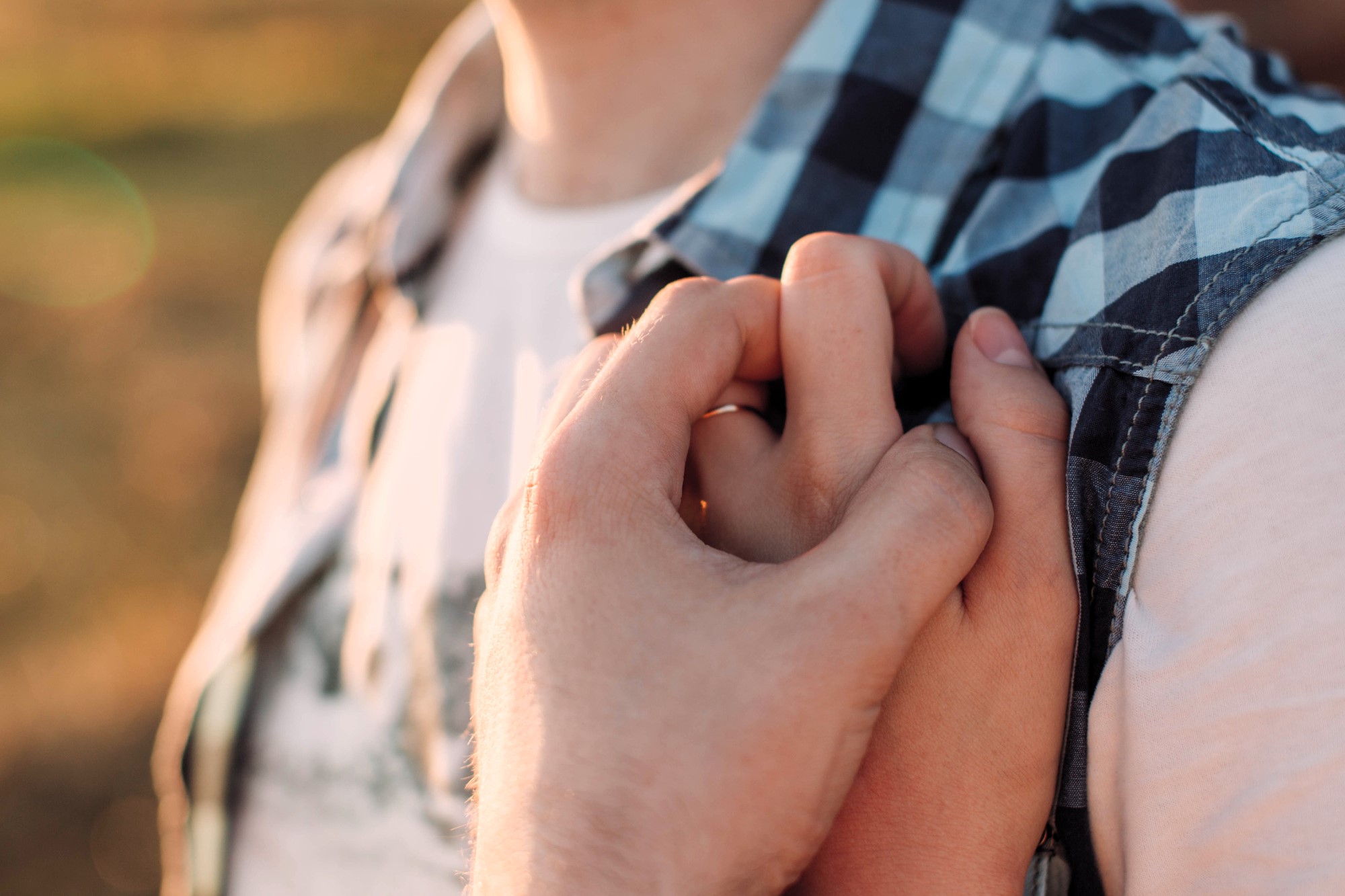 Close-up of two intertwined hands. One hand holds the other's shoulder, over a blue and white checkered shirt, signifying support or connection. The background is softly blurred with warm, natural lighting.