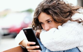 A woman with curly hair, wearing a white sweater, is sitting with her arms resting on her knees. She is holding a smartphone and looking at it intently. The background is blurred, suggesting an outdoor setting.