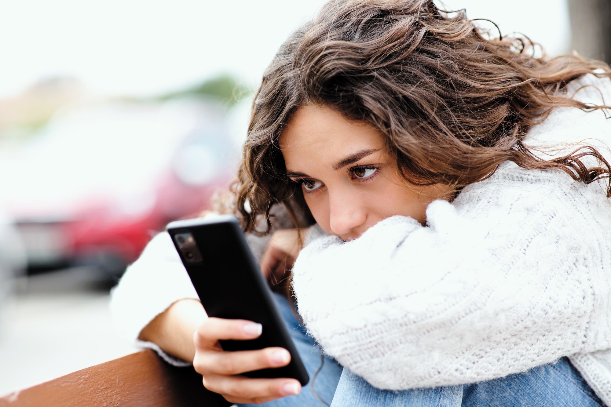 A woman with curly hair, wearing a white sweater, is sitting with her arms resting on her knees. She is holding a smartphone and looking at it intently. The background is blurred, suggesting an outdoor setting.